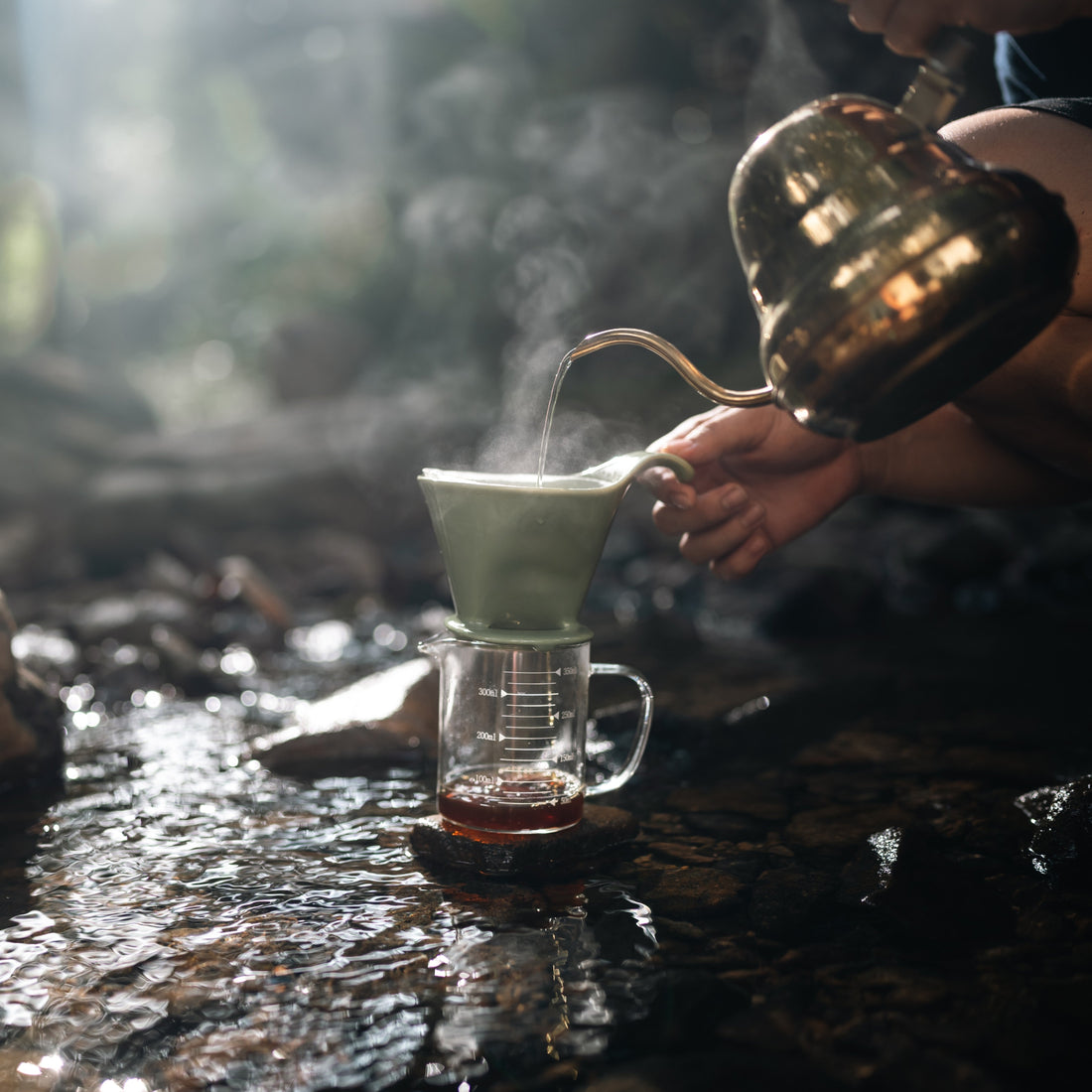 Gooseneck Kettle pouring steaming water into a pour-over dripper outside in stones and stream