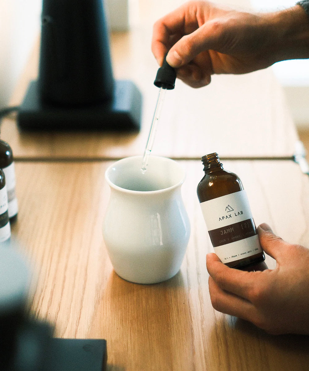 APAX Lab Jamm water mineral bottle held in one hand with dropper in the other hand being dripped into a white tasting cup - all on a wood countertop with a base of a black lamp in the background of the image