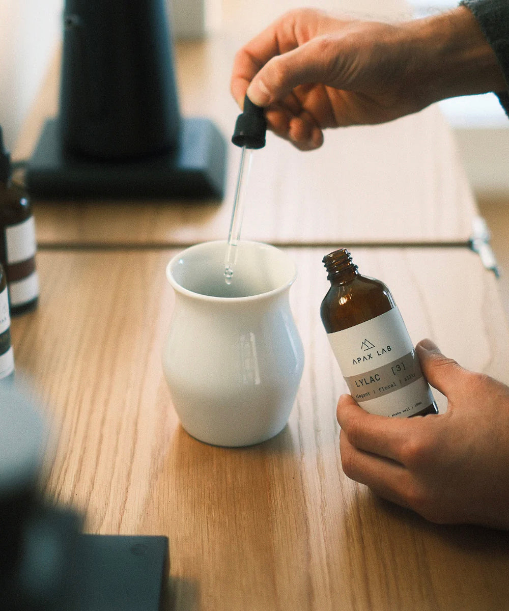 APAX Lab Lylac water mineral front view of bottle being held in one hand with dropper in the other dripping water minerals into a white tasting cup sitting on a wooden counter with other mineral bottles in the background
