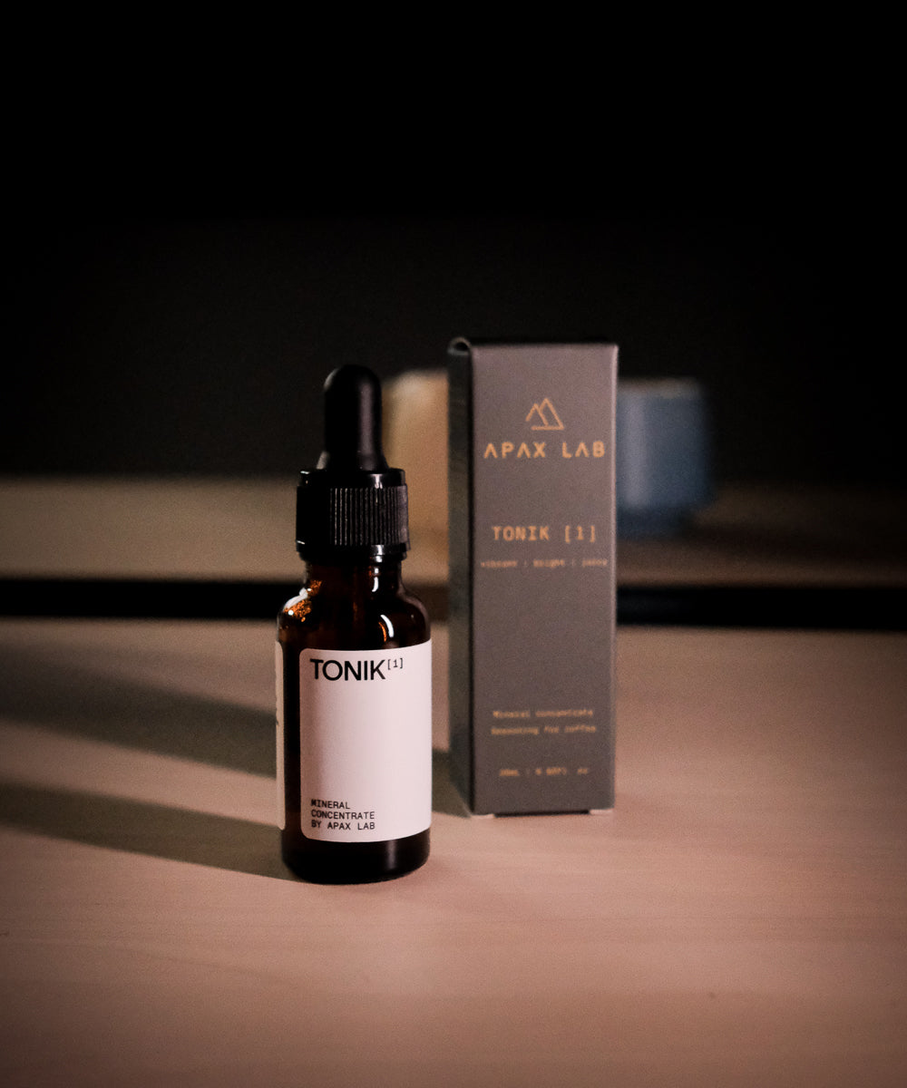 APAX Lab Tonik water mineral front view of bottle with front view of box bottle comes in sitting next to it.  Both on brown table with 2 mugs in the distant background