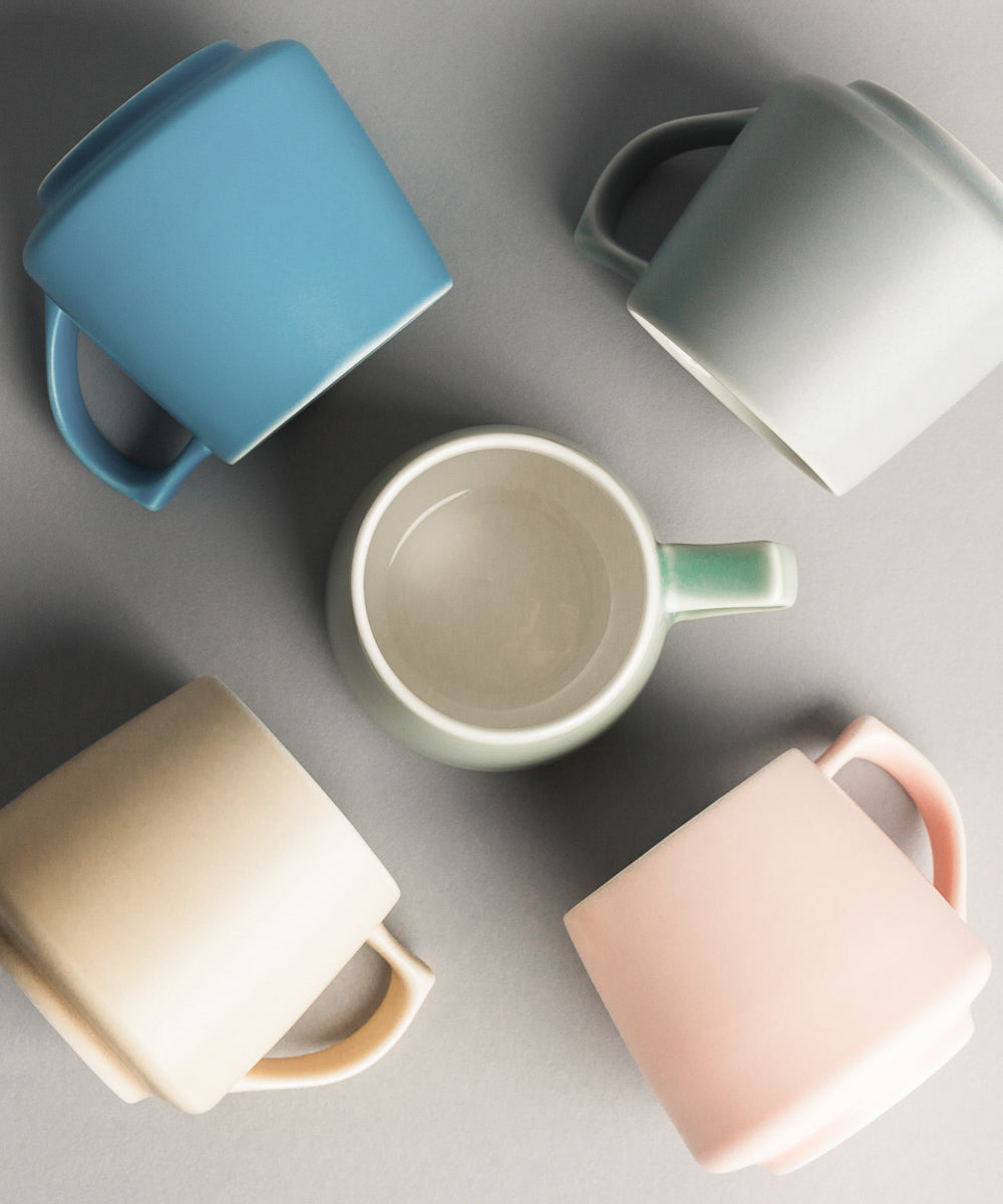 Five ceramic origami mugs in blue, green, beige, and pink on a gray background
