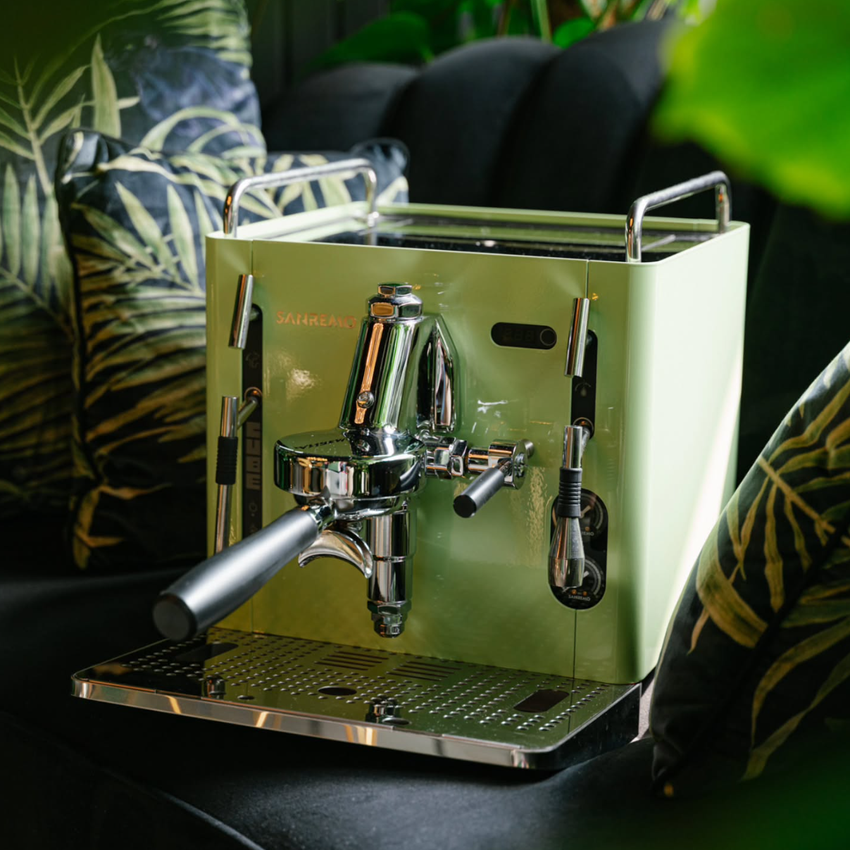 Sanremo CUBE A R  matcha Green home espresso machine with chrome details on a dark surface, surrounded by leafy plants.