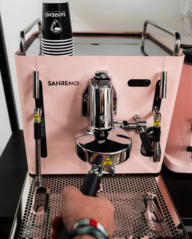 Sanremo CUBE A R  candy pink rose home espresso machine with a hand holding a portafilter and Sanremo paper cups stacked on top of it.